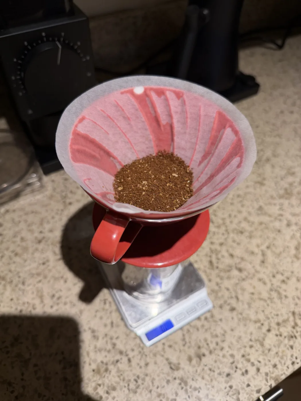 A red Hario V60 sitting on a scale with a bed of dry grounds in a paper filter, a Fellow Ode grinder behind it on the counter.