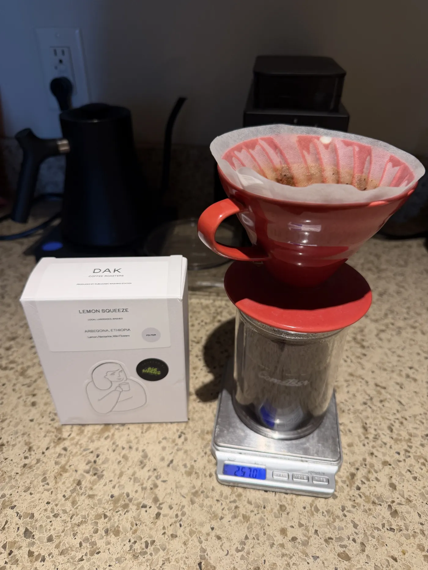The blooming coffee bed in a red Hario V60 on a scale, the DAK Lemon Squeeze box alongside.