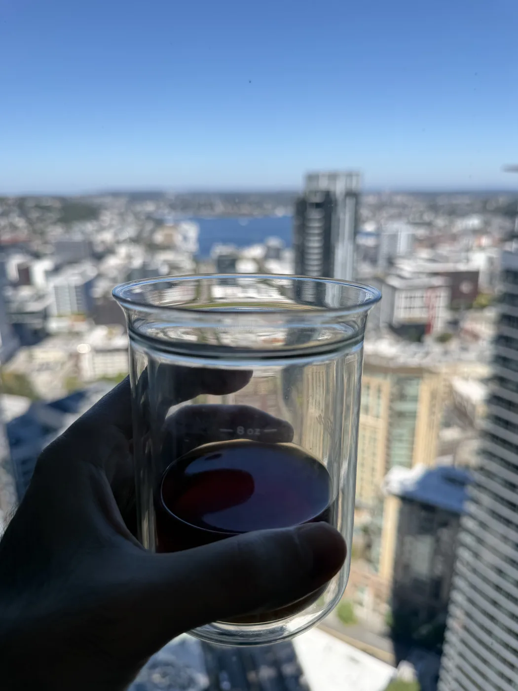 A glass cup of pale amber filter coffee held up against a sunlit view over downtown Seattle, with Lake Union and high-rise towers below.
