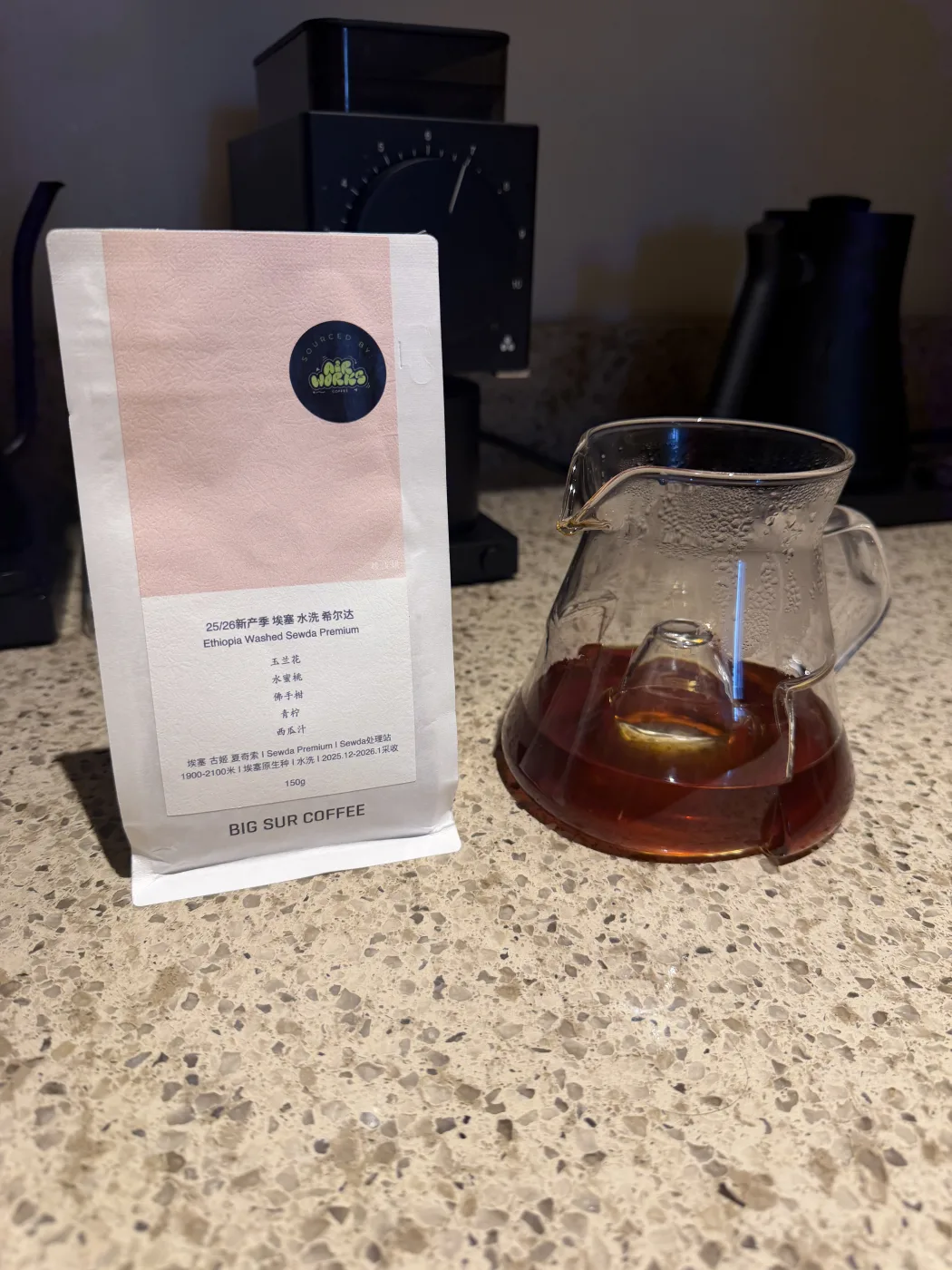 The Big Sur Coffee bag standing beside a glass carafe of brewed coffee with a raised cone in the center of its base, on a kitchen counter with a grinder and kettle behind.
