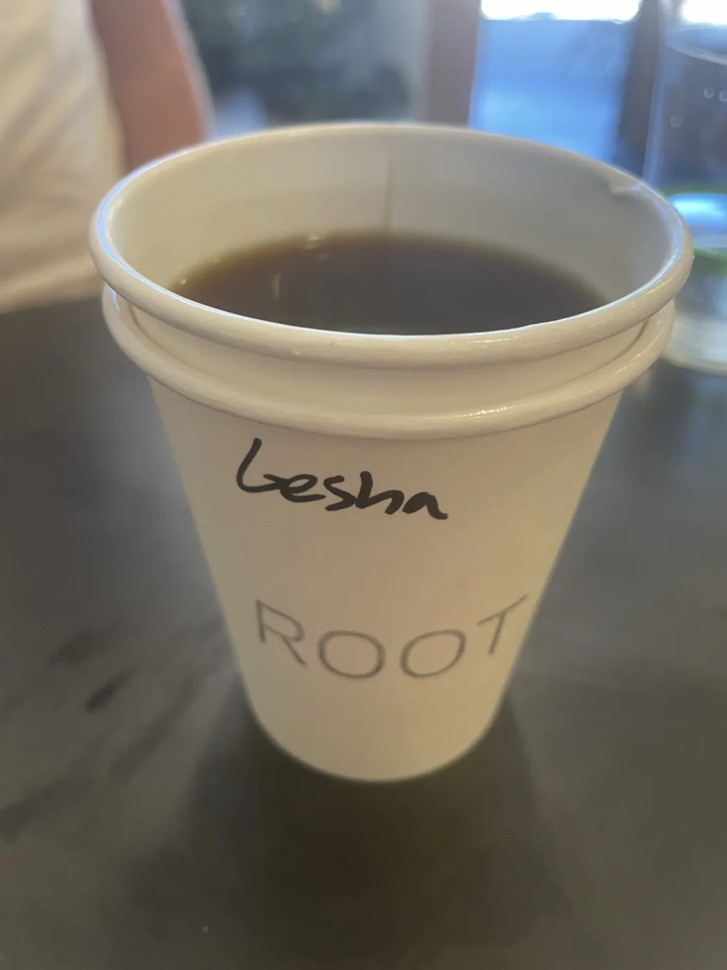 A white ROOT paper cup of black filter coffee on a dark table, with the word Gesha written on the side in marker.