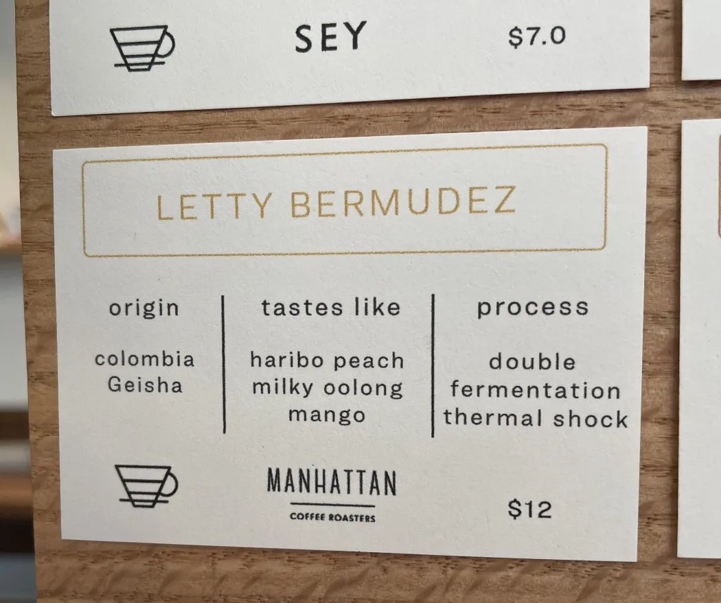 A letterpress menu card at Root reading LETTY BERMUDEZ, origin colombia Geisha, tastes like haribo peach, milky oolong and mango, process double fermentation thermal shock, from Manhattan Coffee Roasters, twelve dollars. A card for Sey at seven dollars sits above it.