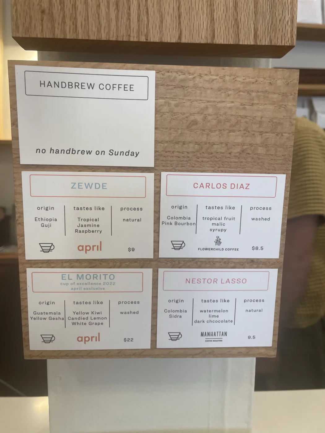 A wooden menu board at Root in Ballard holding five letterpress cards. One reads HANDBREW COFFEE and no handbrew on Sunday. The others list Zewde from April at nine dollars, Carlos Diaz from Flowerchild at 8.50, El Morito from April at twenty-two dollars marked cup of excellence 2022 and april exclusive, and Nestor Lasso from Manhattan at 9.50.