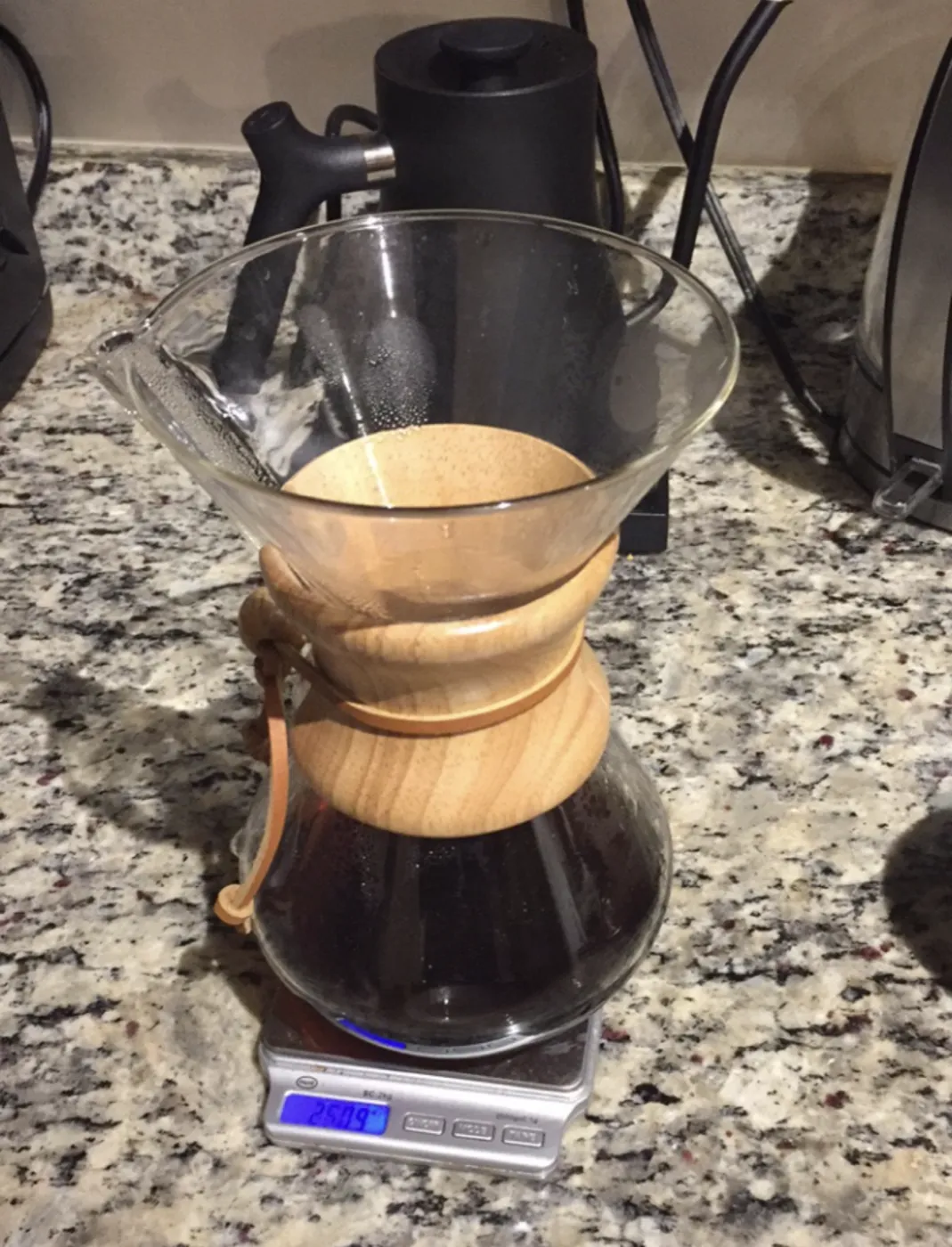 A finished Chemex brew sitting on a scale next to a kettle, the glass base full of dark coffee.