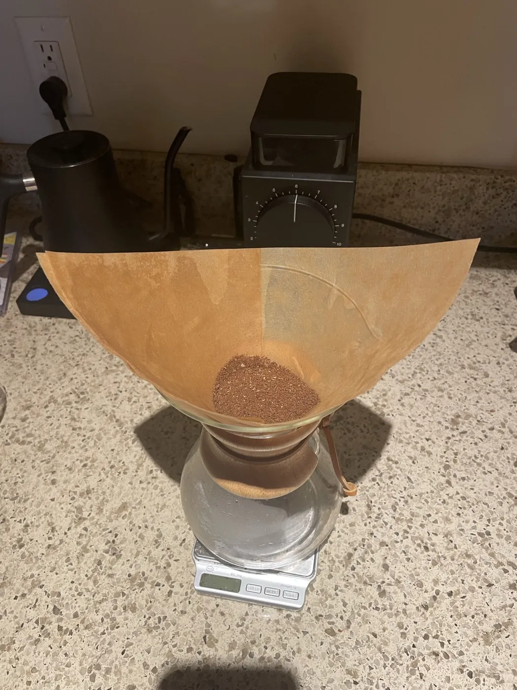 Top-down view of a Chemex with a paper filter open and a dose of ground coffee in the cone, a grinder behind it on the counter.