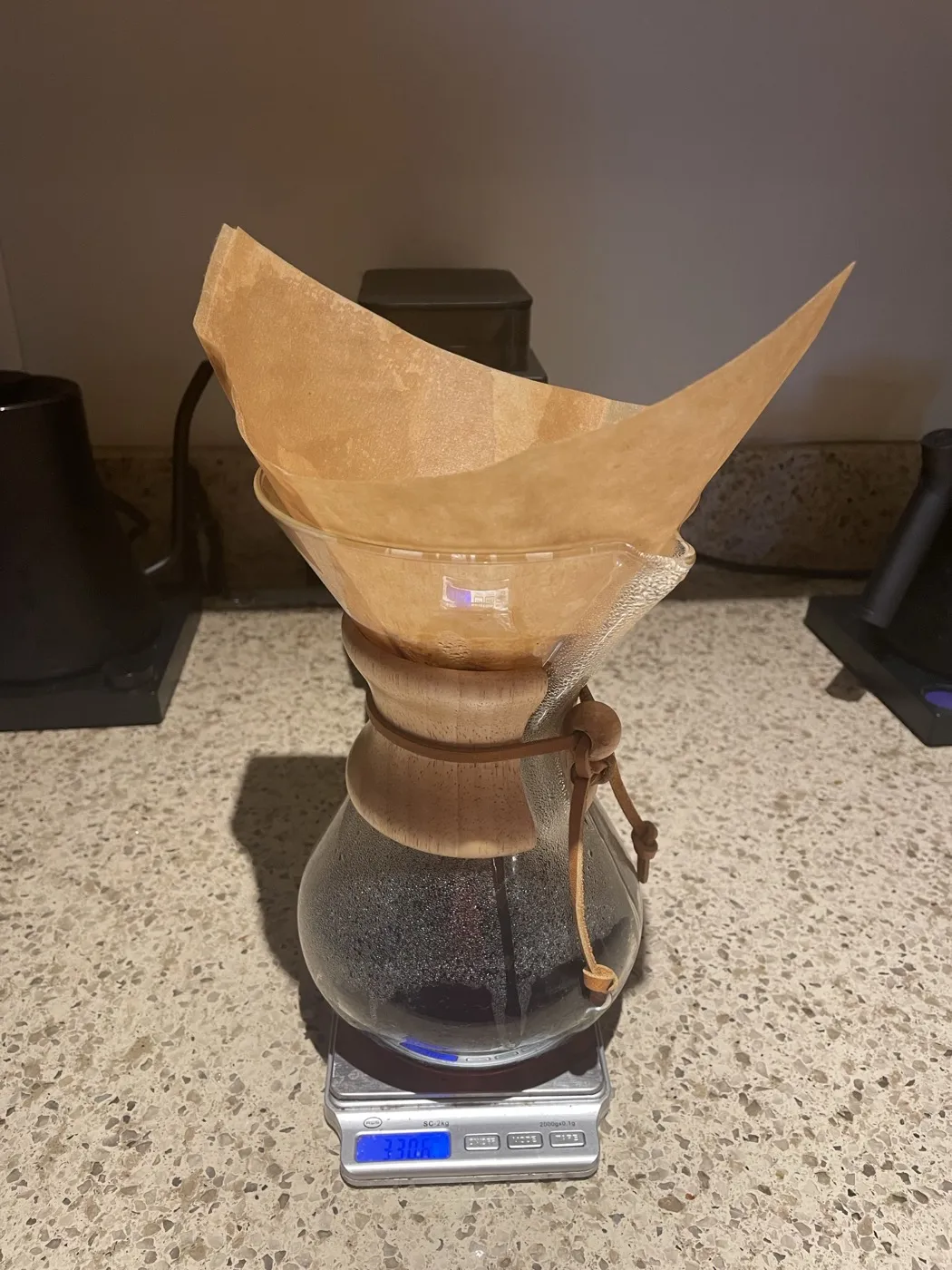 A Chemex on a kitchen scale with its thick tan bonded filter folded open at the top and brewed coffee resting in the glass base.