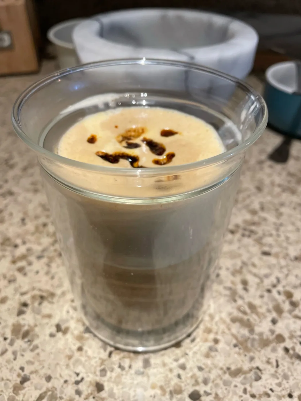 A double-walled glass of latte on a speckled quartz counter, seen from slightly above. Pieces of dark chili crisp sit on top of a pale, intact layer of milk foam, and the espresso and milk layers are visible through the side of the glass.