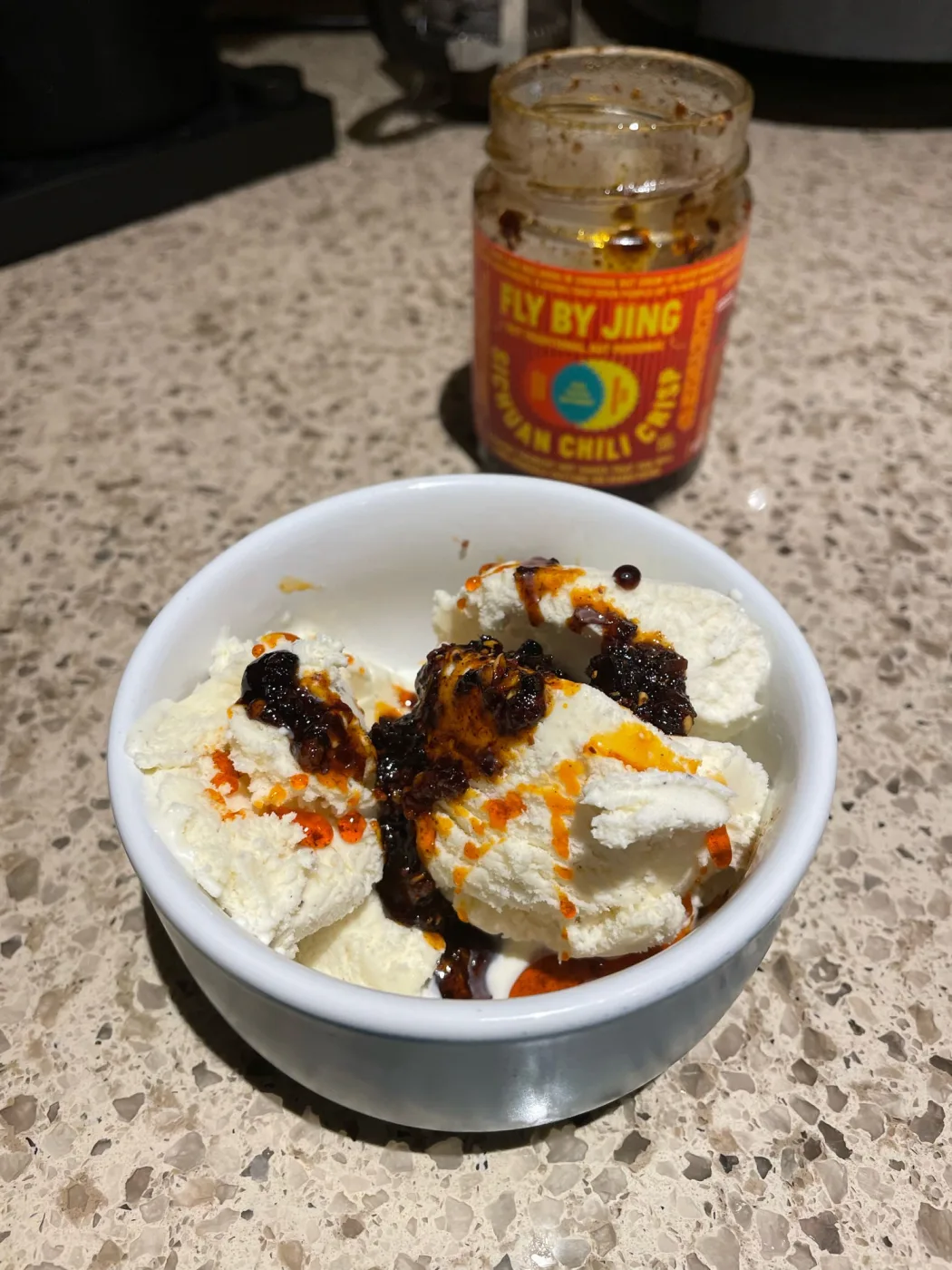 A white ceramic bowl of vanilla ice cream on a speckled quartz counter, with dark chili crisp solids and orange chili oil spooned over the scoops. An open jar of Fly By Jing Sichuan Chili Crisp stands behind it.