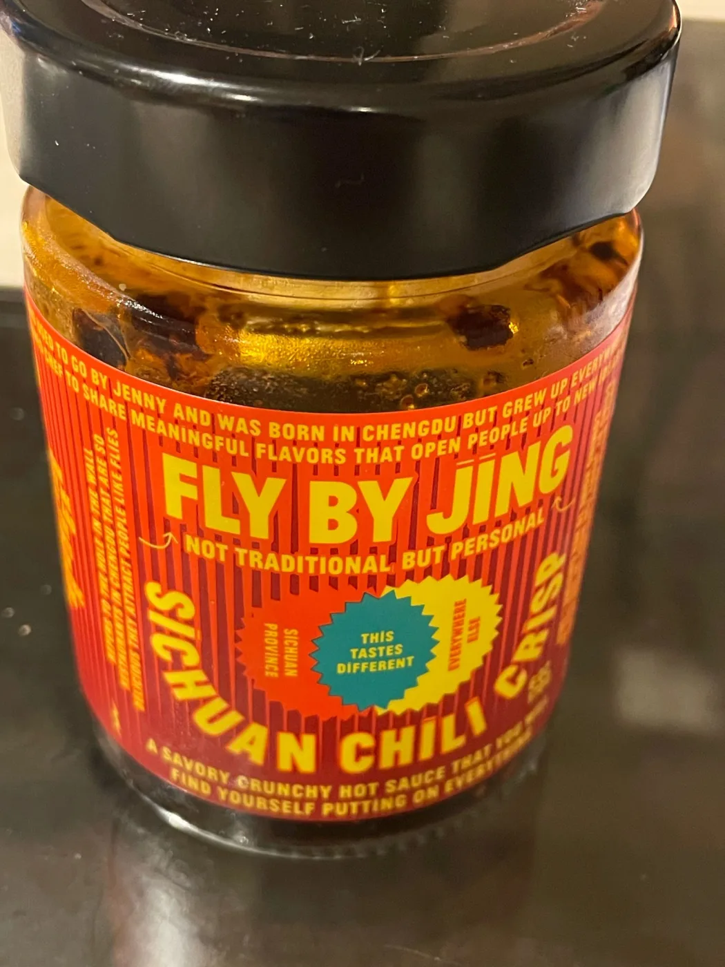 Close-up of a jar of Fly By Jing Sichuan Chili Crisp with a black lid, showing the orange and red label reading FLY BY JING, NOT TRADITIONAL BUT PERSONAL, SICHUAN CHILI CRISP, and a teal starburst reading THIS TASTES DIFFERENT. Chili solids are visible suspended in oil above the label line.