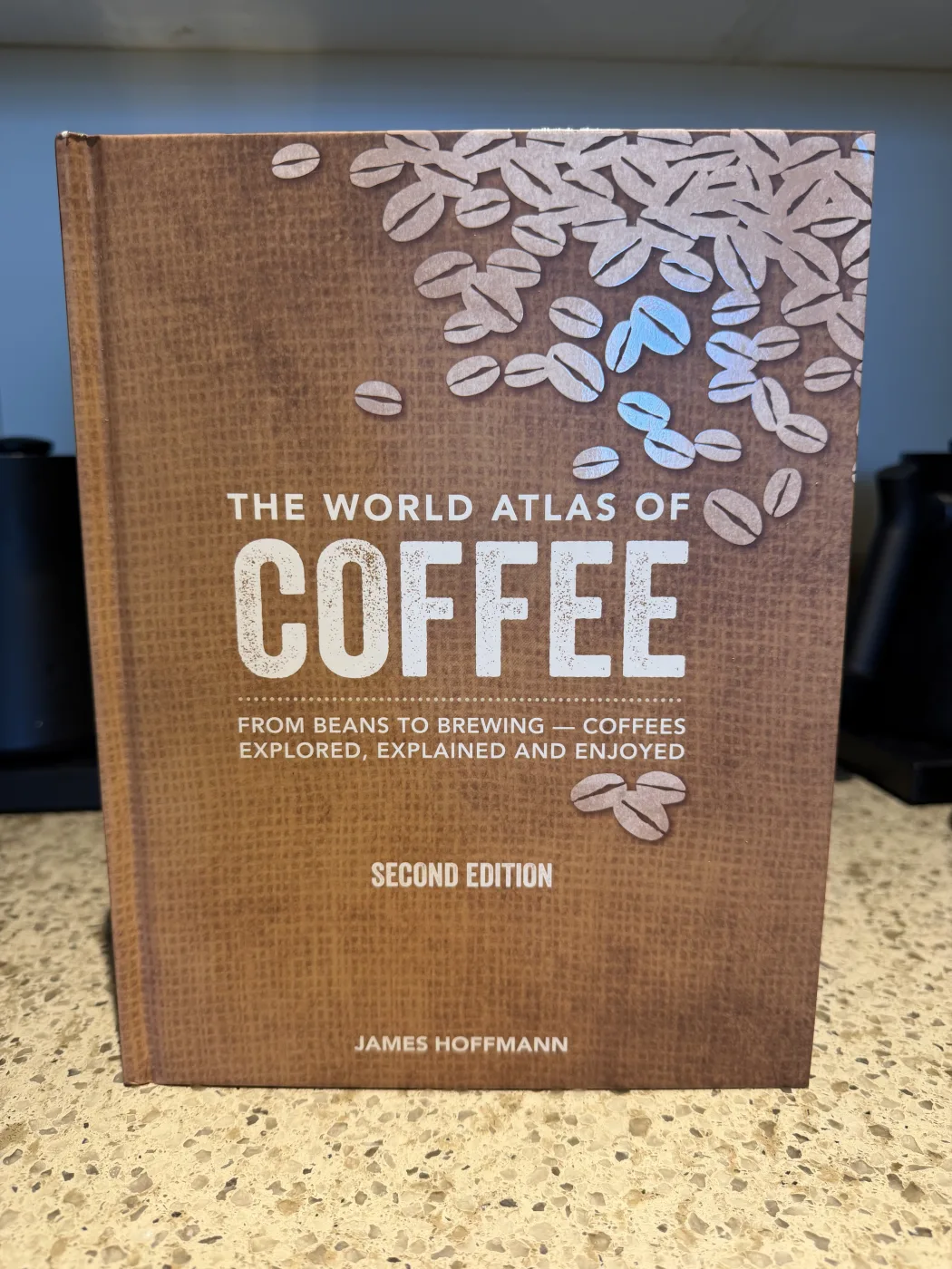 The World Atlas of Coffee, second edition, by James Hoffmann, standing upright on a kitchen counter. The brown cover shows scattered coffee bean illustrations with two beans picked out in blue foil above the title.