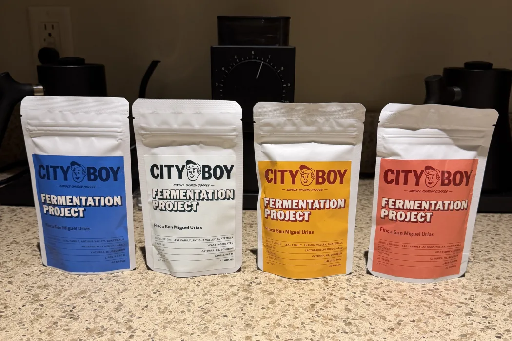 Four City Boy Coffee bags in a row on a speckled countertop, labeled Fermentation Project, Finca San Miguel Urias. From left: blue, white, yellow and orange. A grinder and two gooseneck kettles sit behind them.