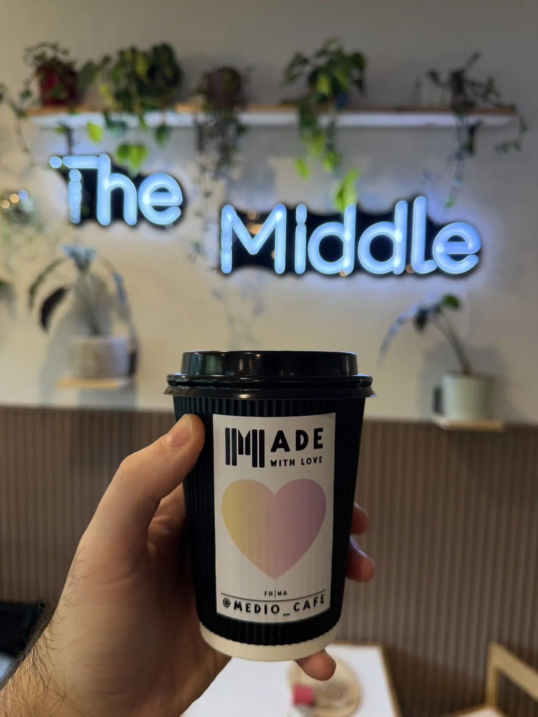 A hand holding a black Medio Cafe takeaway cup with a MADE WITH LOVE label and a pink and yellow heart, in front of a pale blue neon sign reading The Middle, with trailing plants on a shelf behind.