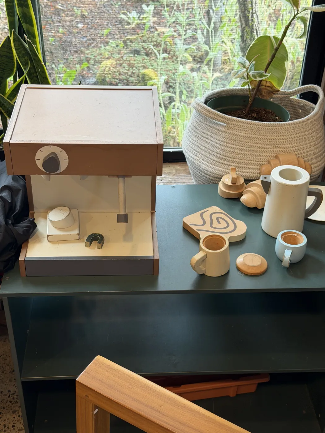 A wooden toy espresso machine on a green table at Medio Cafe, with a control knob, a group head and a small portafilter, beside a wooden kettle, two wooden cups, a wooden croissant and a saucer, in front of a window with plants and a woven basket.