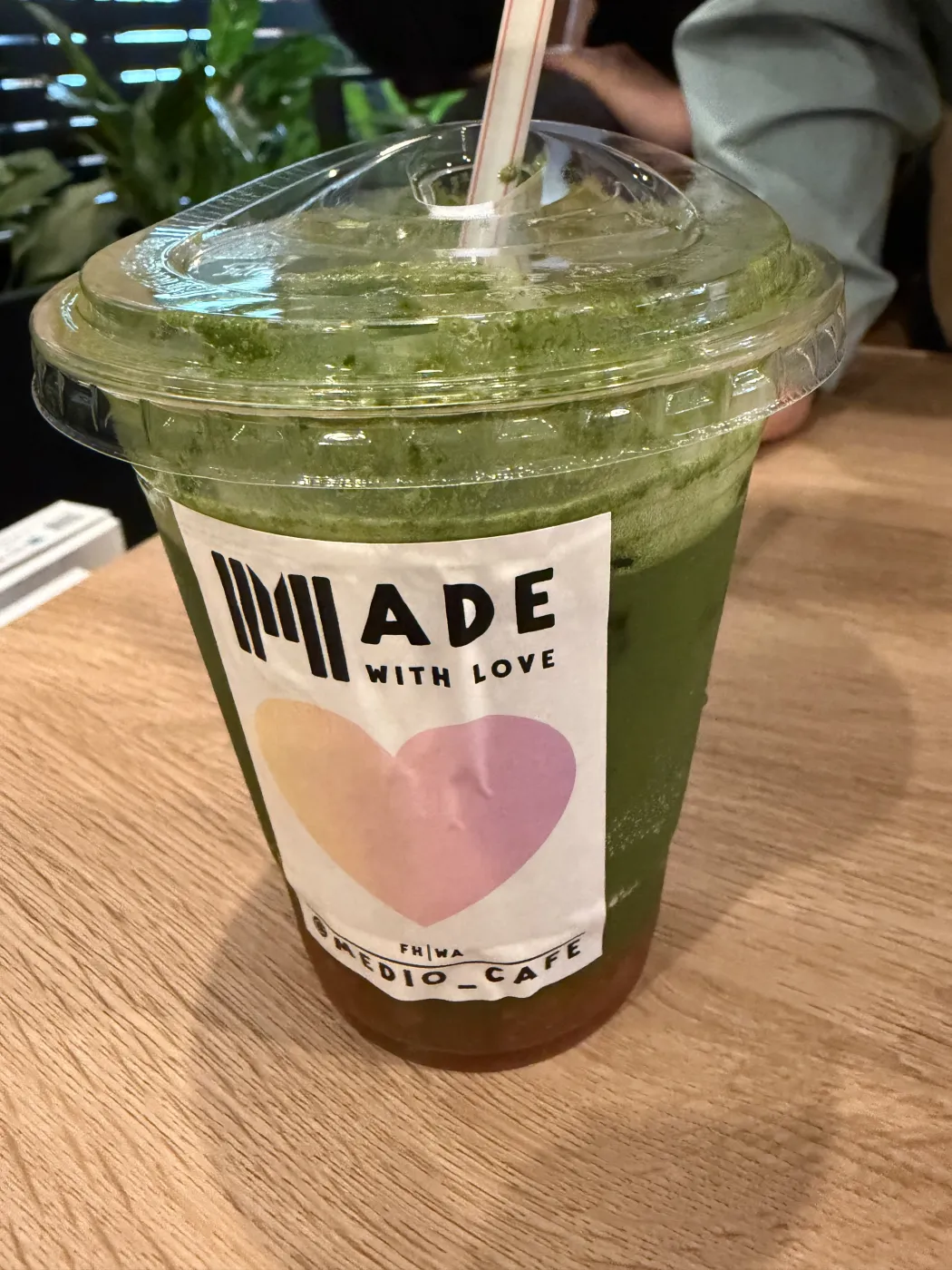 A tall clear plastic cup of sparkling green strawberry matcha over ice with a paper straw, on a light wood table, wearing a Medio Cafe MADE WITH LOVE sticker.