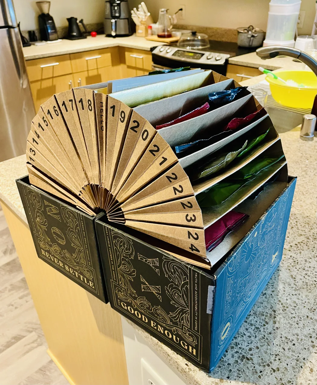 The advent calendar opened on a kitchen counter, with a fan of numbered cardboard tabs from 13 to 24 spread in an arc above the box. Behind the tabs, foil coffee bags in blue, green, red and purple sit in individual card dividers.
