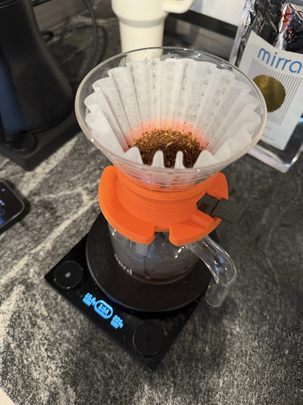 An orange April Hybrid brewer with its flow-control lever, holding a fluted flat-bottom paper filter and a bed of ground coffee, sitting on a glass carafe on a black scale mid-brew, with the Mirra bag behind it.