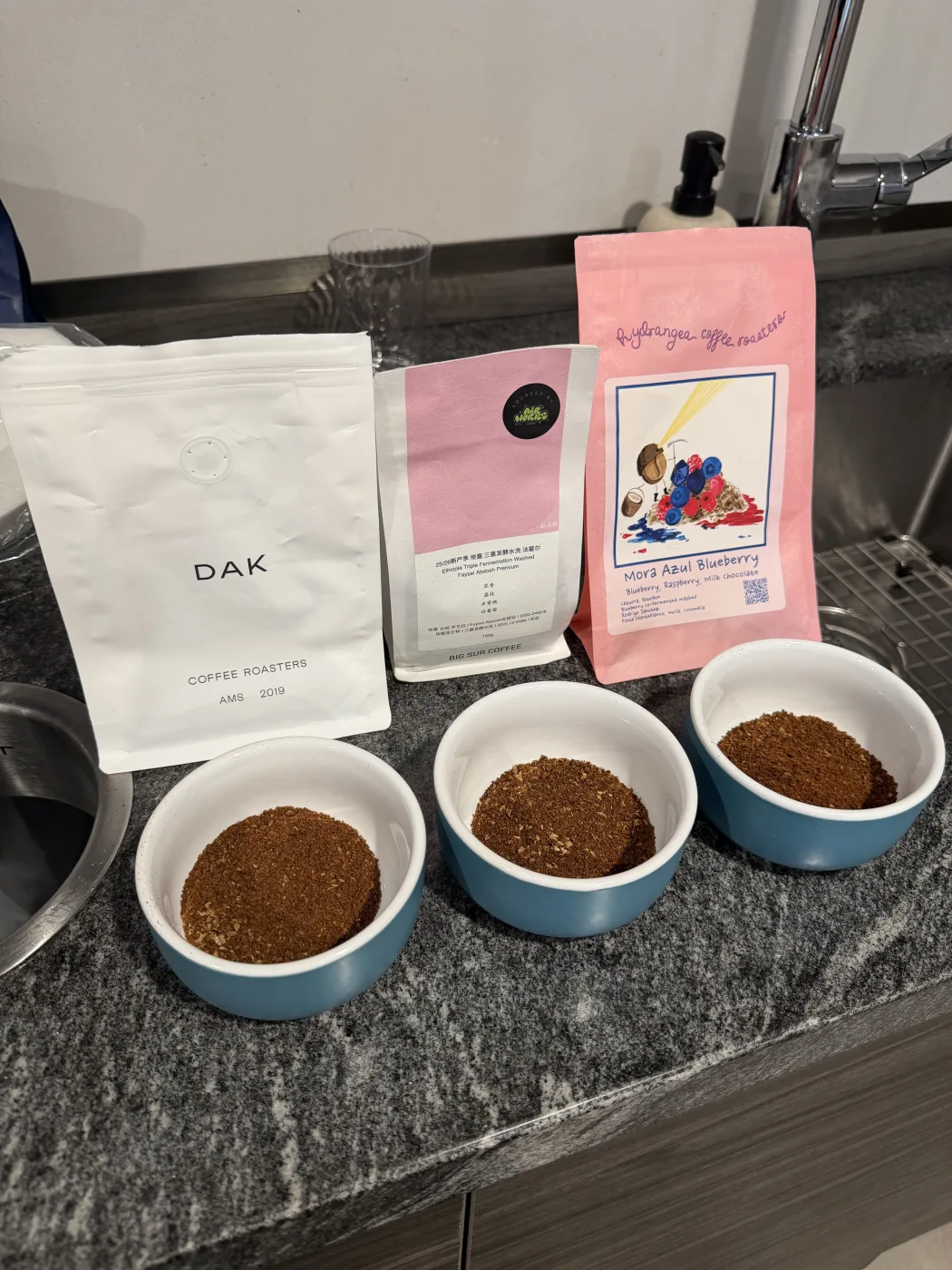 Three blue and white cupping bowls holding dry ground coffee on a granite counter, with their bags laid out alongside: a white DAK Coffee Roasters bag, the Big Sur bag with a sourced-by-Airworks sticker on its pink label, and the pink Hydrangea Mora Azul Blueberry bag with its painted berry artwork.