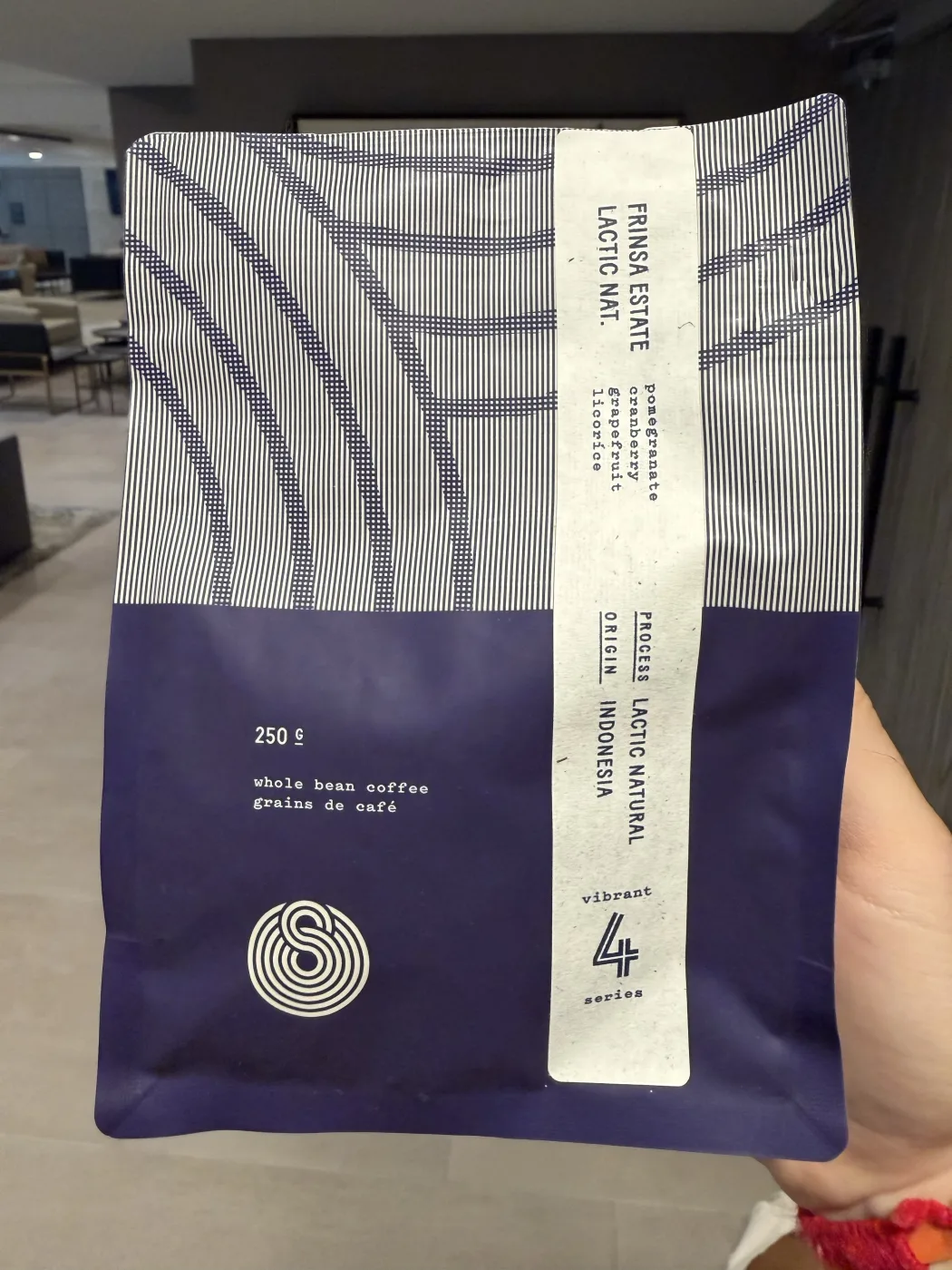A navy and white striped Sought x Found bag held up in one hand. The paper band across it reads Frinsa Estate, Lactic Nat., pomegranate, cranberry, grapefruit, licorice, process lactic natural, origin Indonesia, 250 g whole bean coffee.