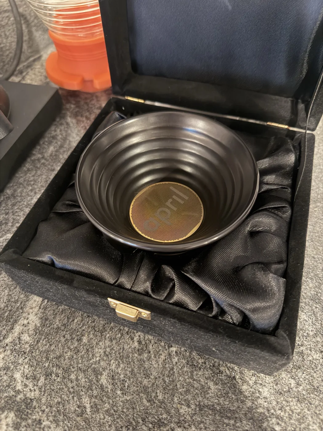 An open black velvet presentation box lined with black satin, sitting on a granite counter and holding a dark ribbed ceramic April brewer with a gold april medallion resting inside it. The orange April Hybrid brewer sits just behind the box.