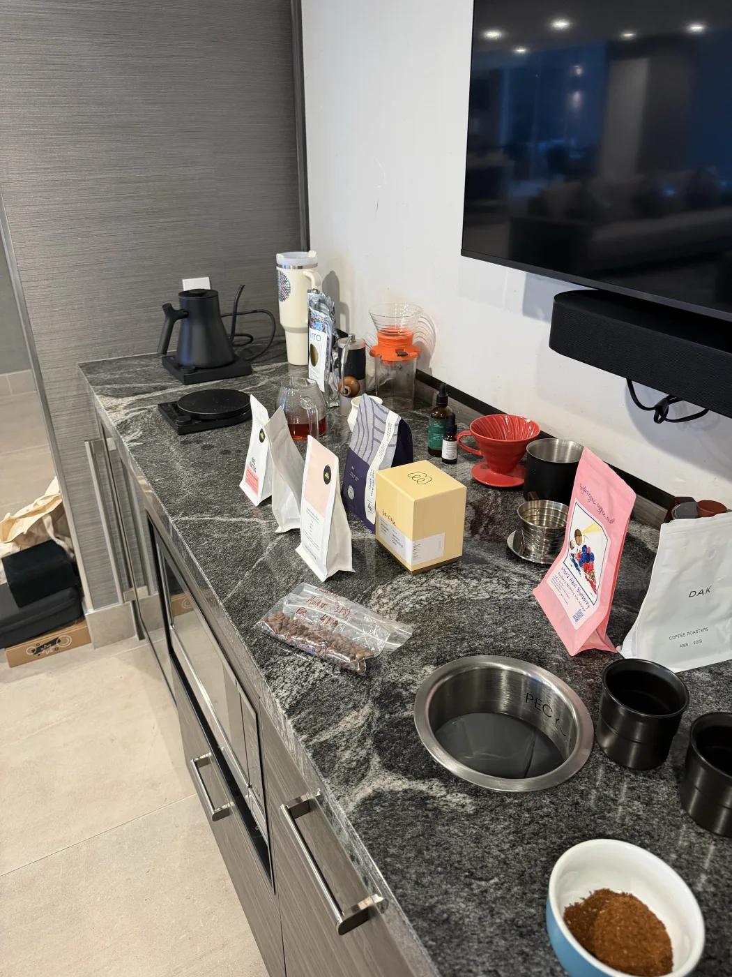 A dark granite kitchen counter seen down its length. A black gooseneck kettle, a hand grinder, and a scale sit at the far end, then a row of coffee bags: two white bags with pink labels, a blue and white striped Sought x Found bag, a pale yellow Monogram box, a red V60 and stainless dosing cups, a pink Hydrangea bag, and a white DAK bag. A clear bag of whole beans and a blue cupping bowl of grounds sit in the foreground.