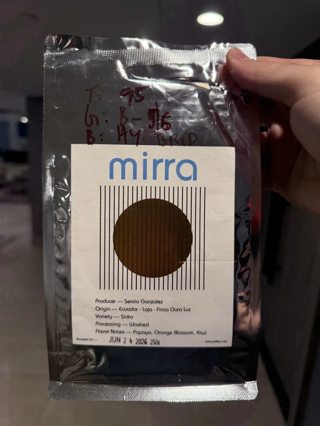 A hand holding a silver Mirra bag in sunlight. The label reads Producer Servio Gonzalez, Origin Ecuador Loja Finca Clara Luz, Variety Sidra, Processing Washed, Flavor Notes Papaya Orange Blossom Kiwi, roasted on Jun 24 2026, with brew parameters written on the foil in red marker.