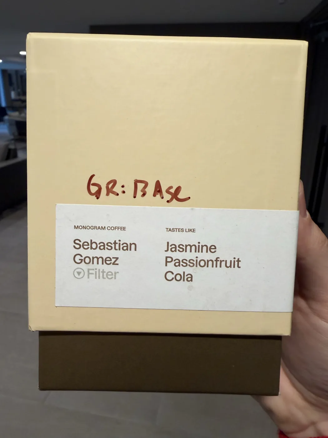 A pale yellow Monogram Coffee box held up in one hand. The band reads Sebastian Gomez, Filter, tastes like Jasmine, Passionfruit, Cola, with a grind setting written on the box in red marker.
