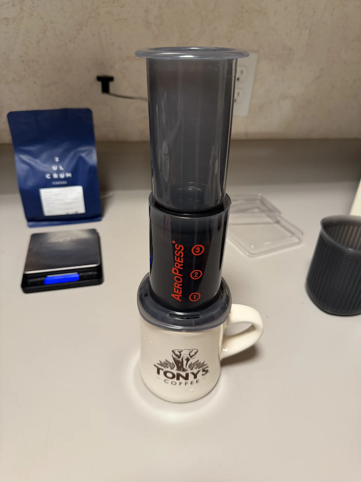An AeroPress Go brewing in the standard orientation on top of a Tony's Coffee mug, chamber full, plunger seated on a hotel room counter.