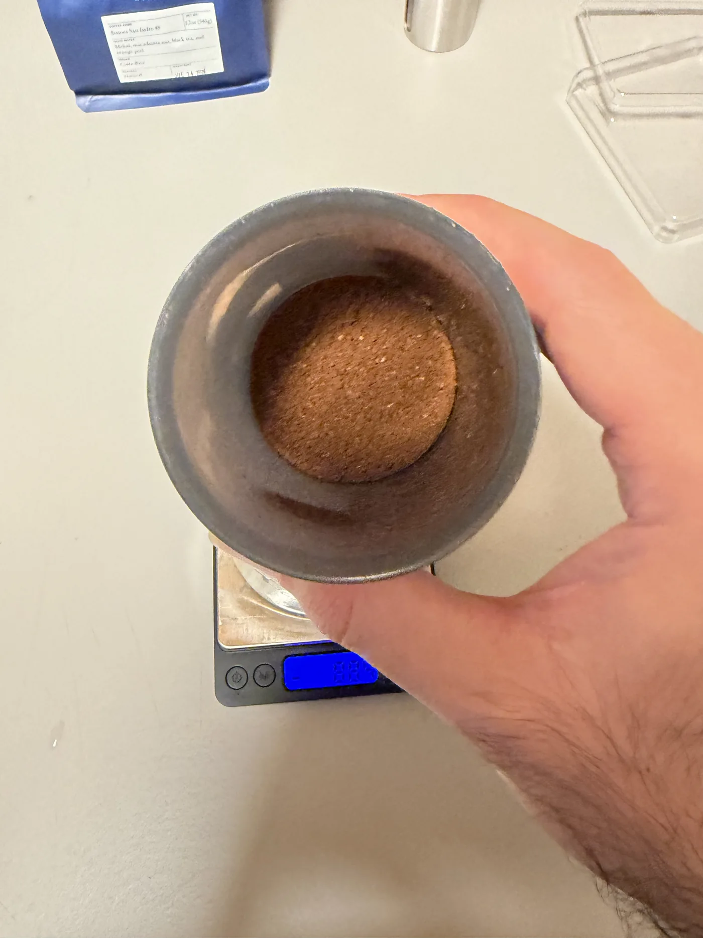 Freshly ground coffee in the catch cup of a stainless steel hand grinder, held over a pocket scale, showing an even medium-fine grind.