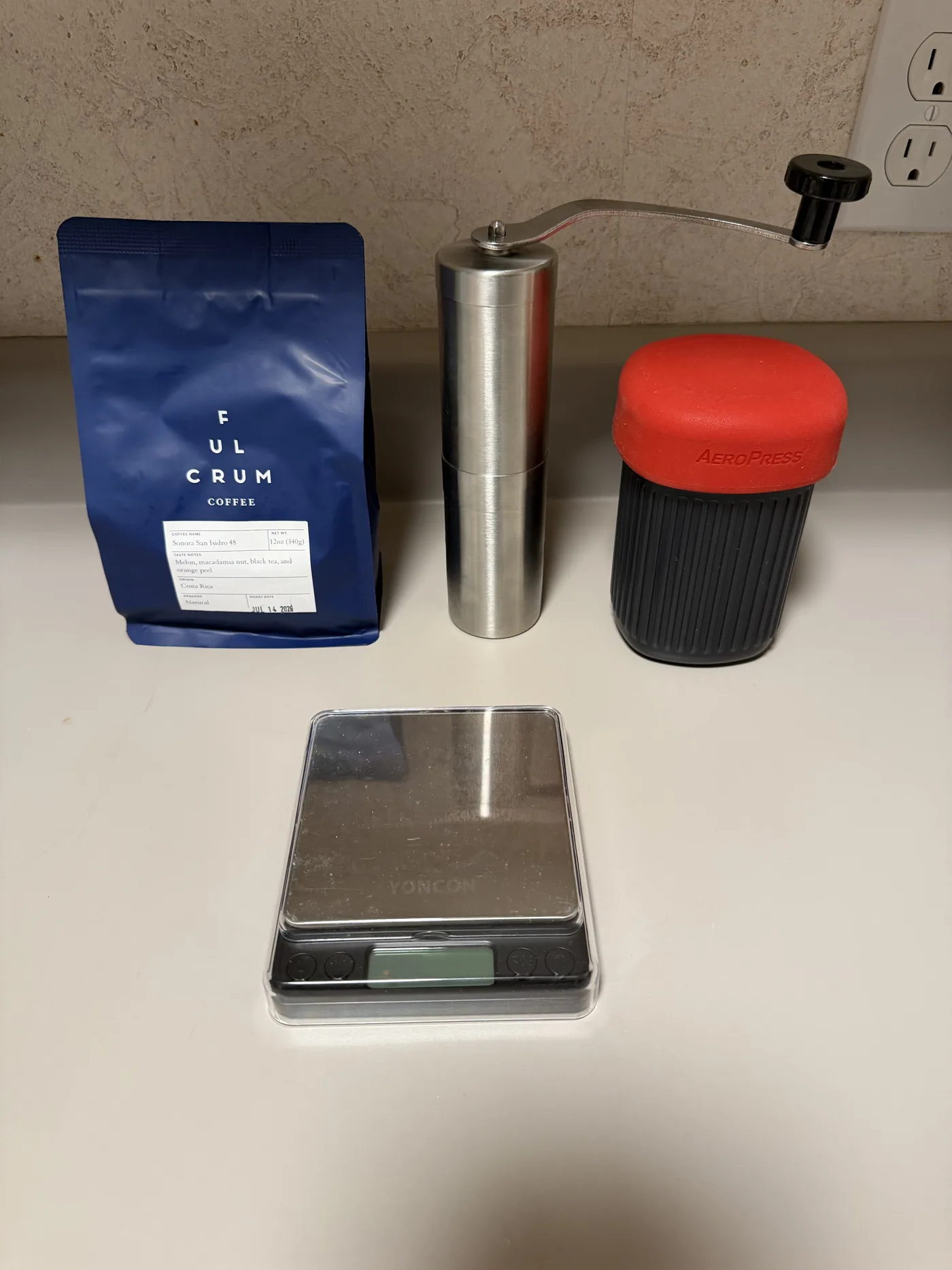 A travel coffee kit laid out on a hotel room counter: a blue Fulcrum Coffee bag labelled Sonora San Isidro 48 from Costa Rica, a stainless steel hand grinder with its crank attached, an AeroPress Go with its red silicone lid on, and a small black digital pocket scale.