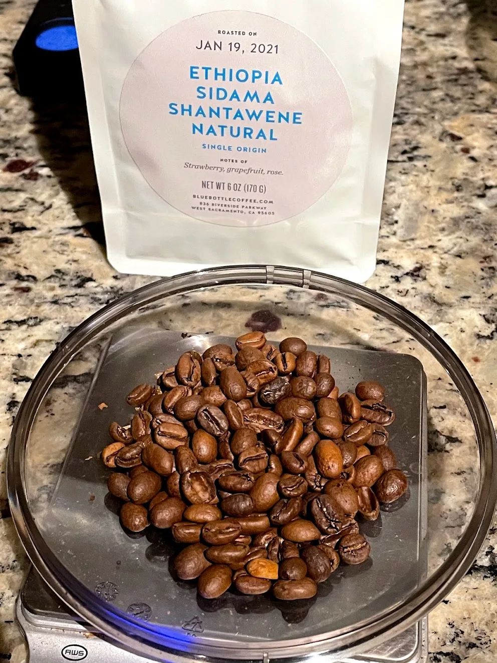 A white Blue Bottle bag labelled ROASTED ON JAN 19, 2021, ETHIOPIA SIDAMA SHANTAWENE NATURAL, notes of strawberry, grapefruit, rose, standing behind a pile of roasted coffee beans on the glass platter of a kitchen scale.