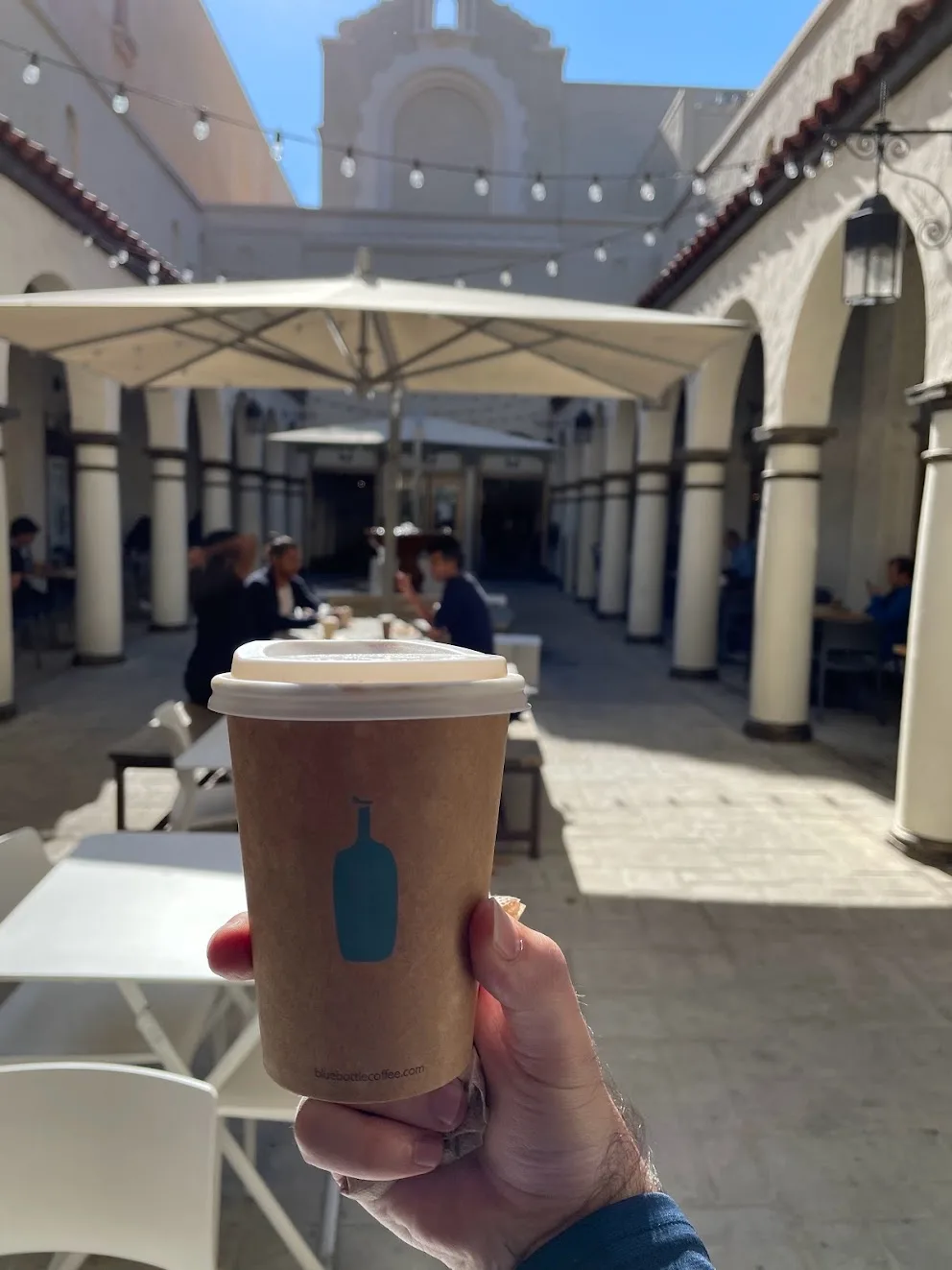 A hand holding a brown Blue Bottle paper cup with a white lid, in a sunlit courtyard with white stucco arches on both sides, red tile roofs, string lights overhead and a church facade at the far end. People sit at tables under a white umbrella.