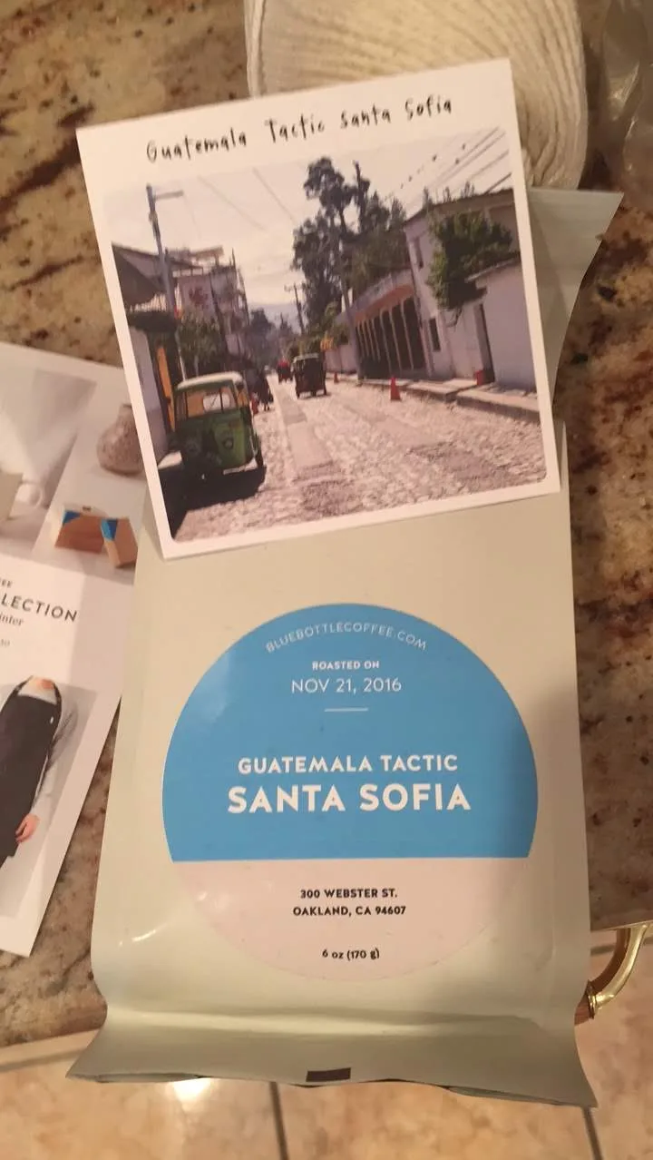 A Blue Bottle bag with a blue and white circular label reading ROASTED ON NOV 21, 2016, GUATEMALA TACTIC SANTA SOFIA, and beneath it the address 300 WEBSTER ST. OAKLAND, CA 94607, net weight 6 oz. A postcard showing a cobbled street with a green tuk-tuk leans against the bag.