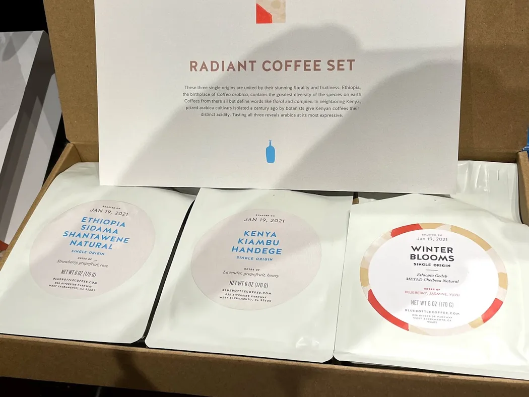 An open cardboard box holding three white Blue Bottle bags, each with a circular label reading ROASTED ON JAN 19, 2021: Ethiopia Sidama Shantawene Natural with notes of strawberry, grapefruit and rose; Kenya Kiambu Handege with lavender, grapefruit and honey; and Winter Blooms, an Ethiopia Gedeb METAD Chelbesa Natural, with blueberry, jasmine and yuzu. A printed card behind them reads RADIANT COFFEE SET.