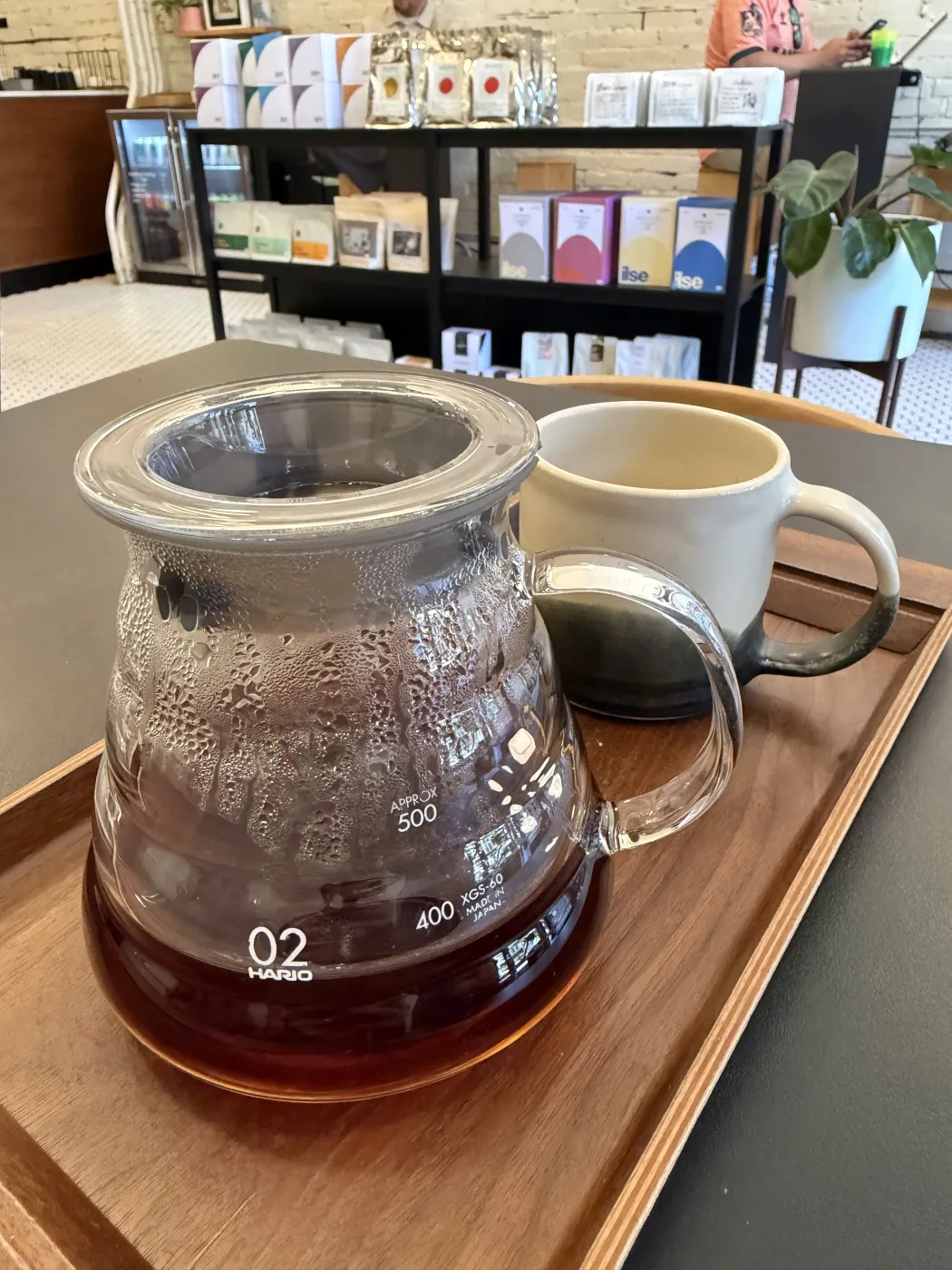 A Hario glass carafe of brewed coffee beside a cream and dark-glazed ceramic mug on a wooden serving tray at Homage Coffee, with bags of coffee on shelves blurred behind.