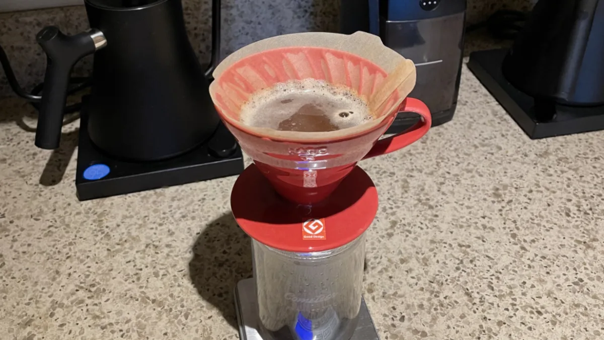 A red Hario V60 blooming over a glass server on a digital scale, with a Baratza Encore grinder and a gooseneck kettle on the counter behind it.